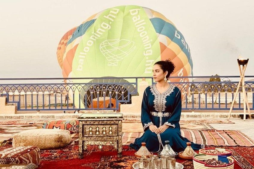 A person sitting on a carpeted area with a traditional chair and a table with a teapot, in front of a colorful hot air balloon. 1