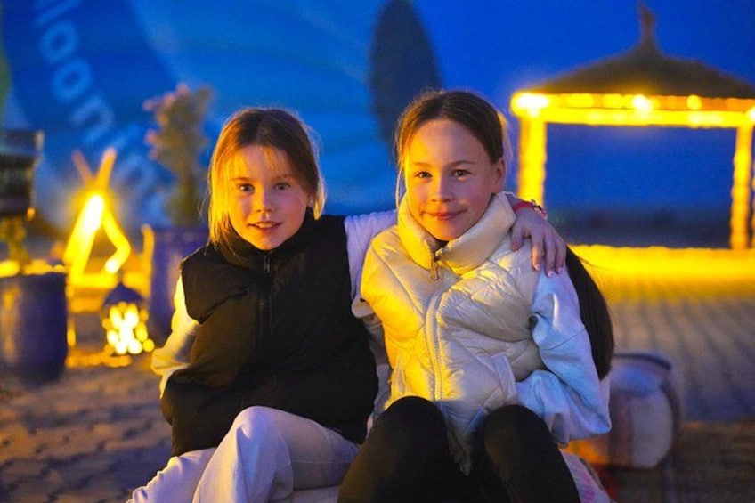 Two children sitting on a wooden deck at night with a gazebo and a campfire in the background. 5