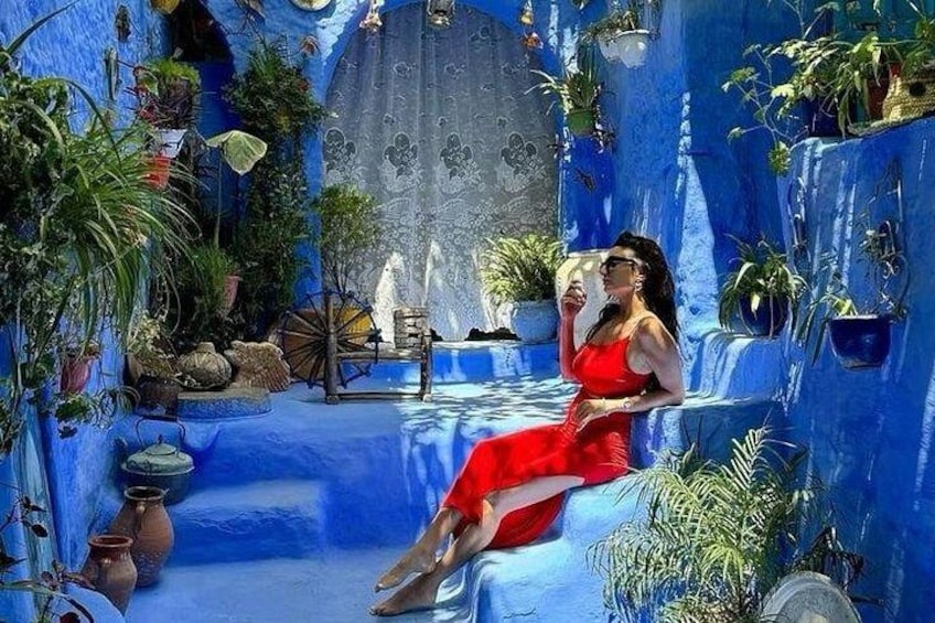 A woman in a red dress sitting on a blue wall with plants and a fountain. 1