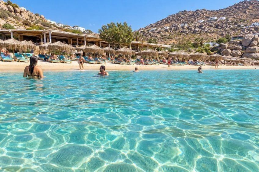 A clear blue pool with people swimming and sunbathing on the beach, surrounded by thatched umbrellas and a mountainous backdrop. 3
