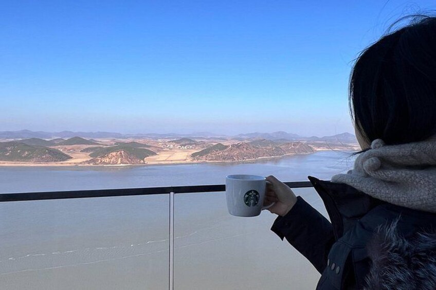 Eine Person hält eine Starbucks-Tasse in der Hand und blickt auf eine Küstenlandschaft mit Hügeln und einem Gewässer. 2