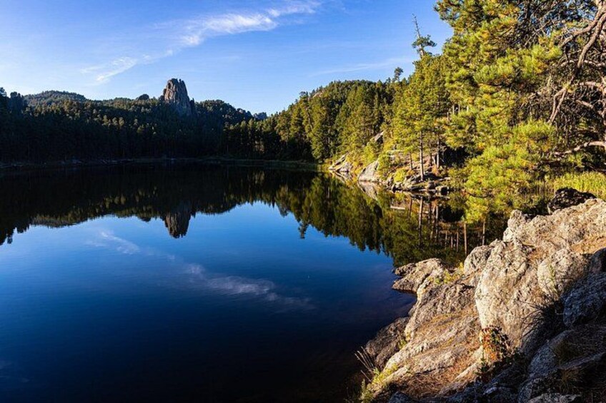 A calm lake with a rocky shoreline, dense forest, and a prominent rock formation in the distance. 2
