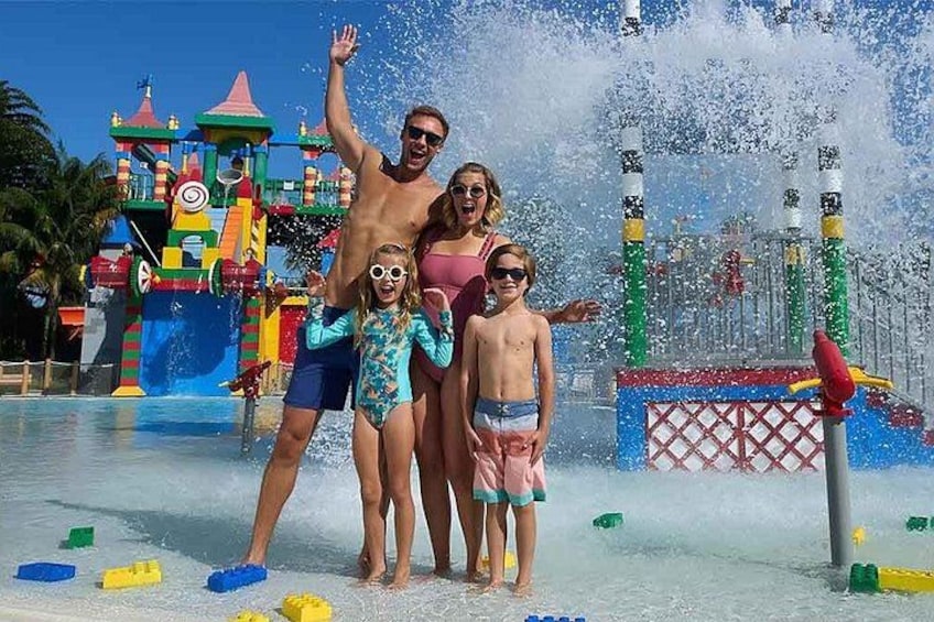 A family at a water park with a colorful play structure and water jets. 1