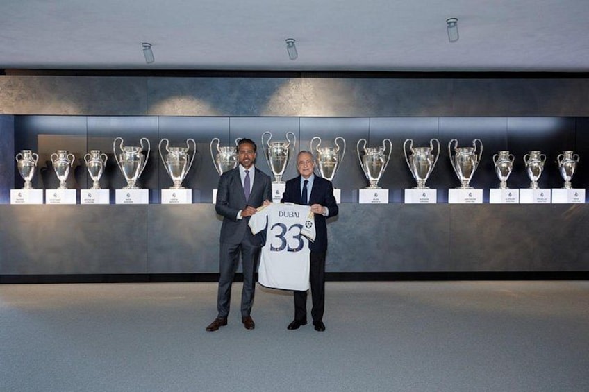 Two men standing in front of a wall displaying trophies. One man holds a jersey with the number 33. 4
