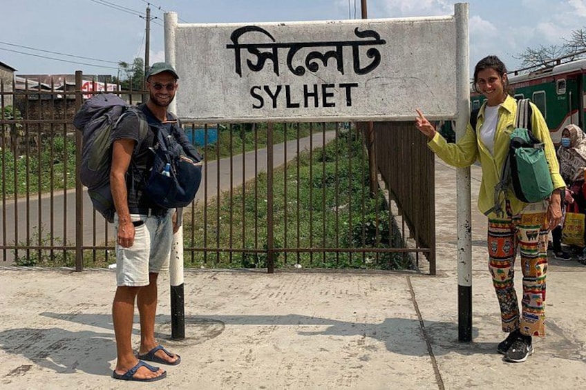 Two people standing in front of a sign with the text 'SYL HET' and a train in the background. 4