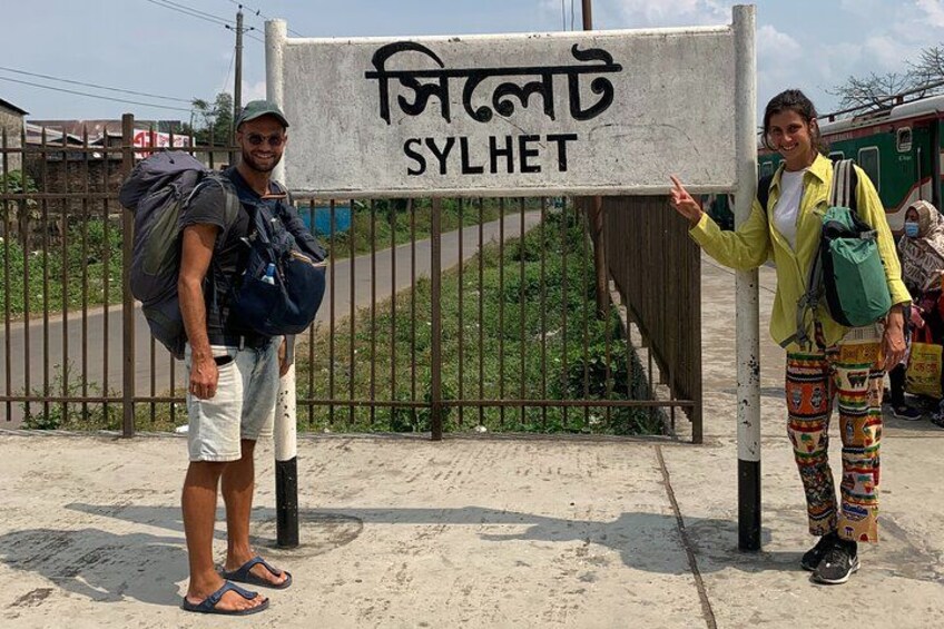 Two people standing in front of a sign with the text 'SYL HET' and a train in the background. 3