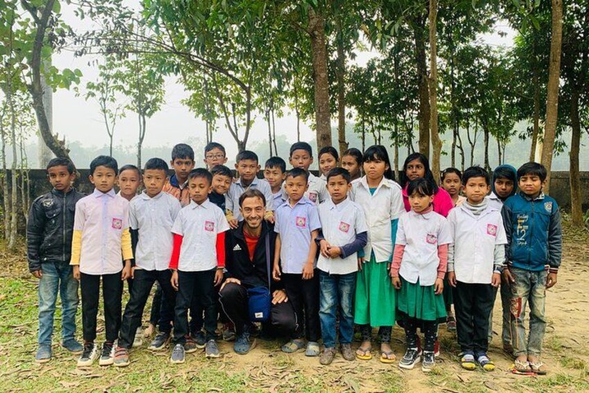 A group of children and adults outdoors, some wearing school uniforms, standing on a dirt ground with trees in the background. 5