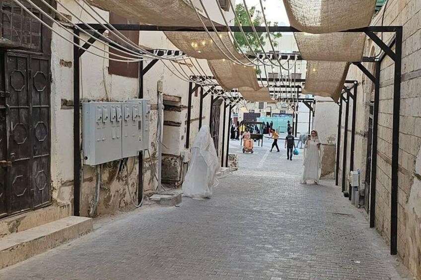 A narrow alleyway with buildings on either side, a covered walkway overhead, and a few people in the distance. 4