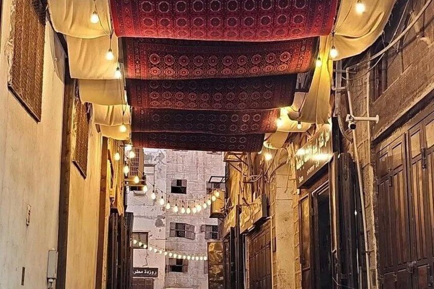 A narrow alley with patterned fabric hanging overhead, string lights, and a building with a sign. 3