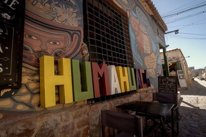 A colorful mural with the word 'HUMAHUALE' on a building facade, a café sign, and a rustic stone wall. 5