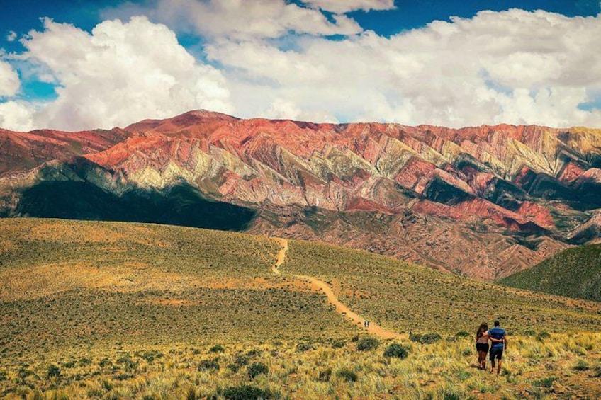 A hiker on a trail with red and green mountains in the background. 3