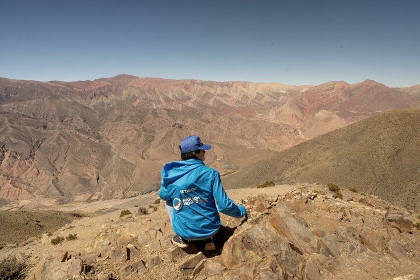 A person in a blue jacket sitting on a rocky outcrop overlooking a mountainous landscape. 3