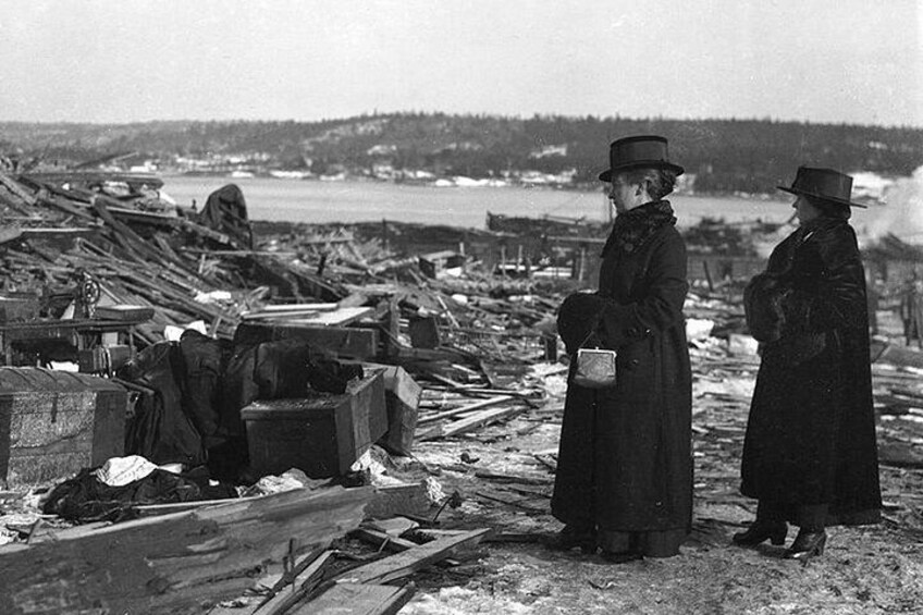 A devastated landscape with debris and wreckage, two women in long coats and hats observing the scene. 4