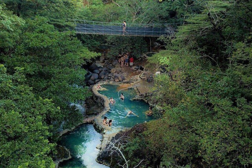Una fuente termal natural rodeada de un bosque denso, un puente de madera y gente disfrutando del agua. 2