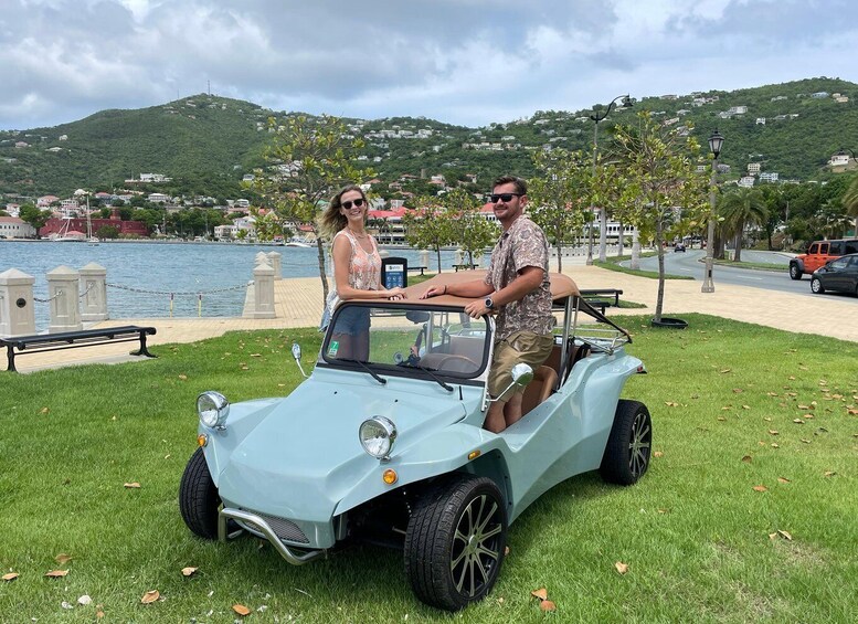 A light blue convertible car parked on grass near a waterfront with a backdrop of hills and buildings. 1