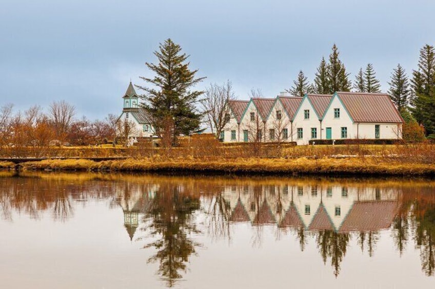 A row of white houses with green trim, a church with a steeple, and a calm lake reflecting the buildings. 2