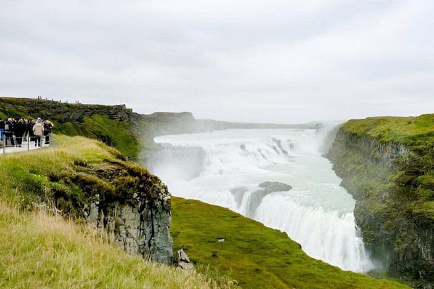 A waterfall cascading down a cliff, surrounded by grassy terrain and a viewing platform with people. 2