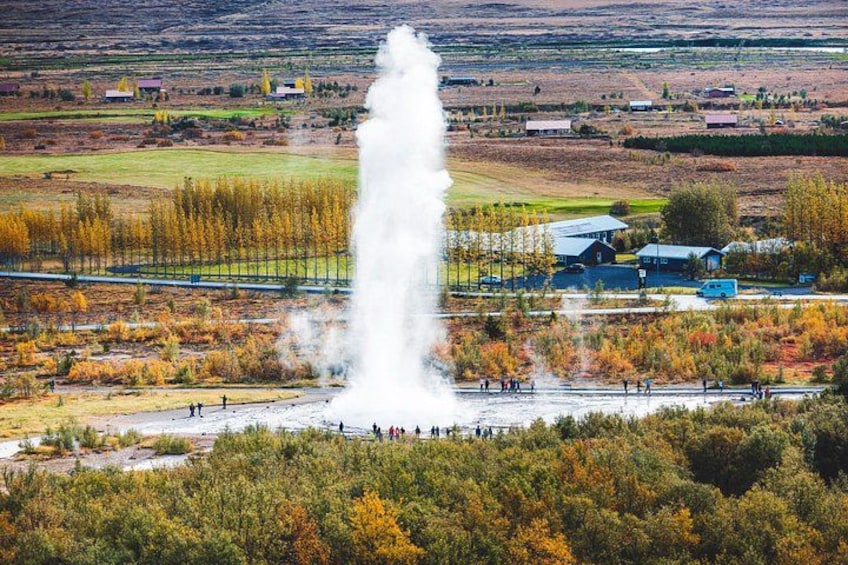 A geyser with people observing it, surrounded by autumn trees and a rural landscape with buildings in the background. 3