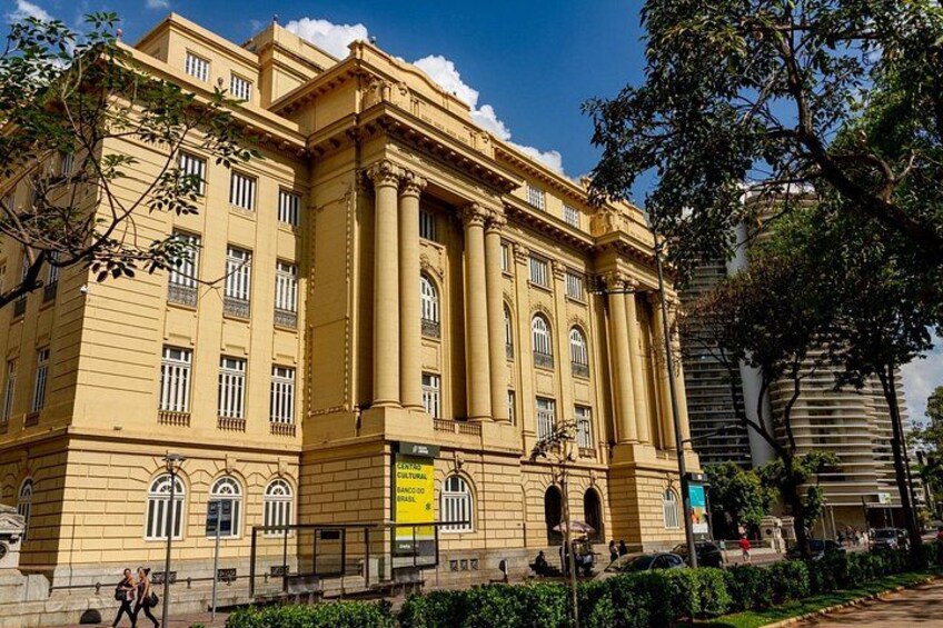 A large yellow building with columns and arched windows, surrounded by greenery and a clear sky. 2