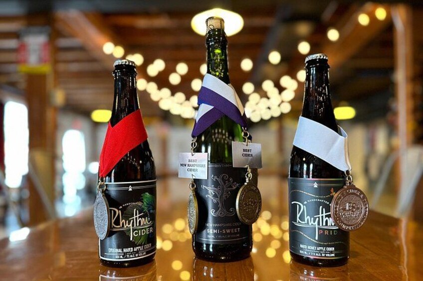 Three bottles of Rhythmic Cider on a reflective surface with a blurred background of lights. 3