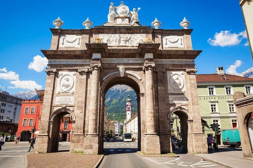 A historic stone archway with intricate carvings and statues, leading to a town square with buildings and a mountain in the background. 2