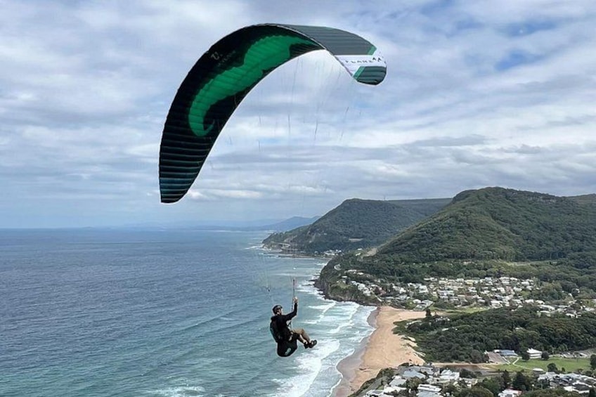 Una persona practicando parapente sobre una zona costera con playa y colinas al fondo. 1