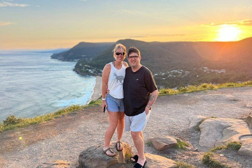 Two people standing on a rocky outcrop overlooking a coastal landscape with a sunset in the background. 2