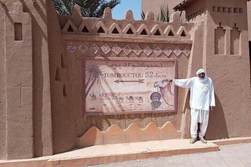 A man standing in front of a wall with a sign that reads 'Tombouctou 52 Joutes' and includes illustrations of camels and people. 5