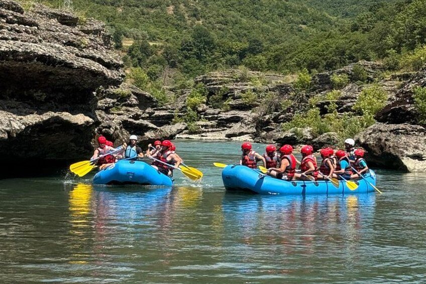 Two blue inflatable rafts with people paddling on a calm river surrounded by rocky terrain. 2