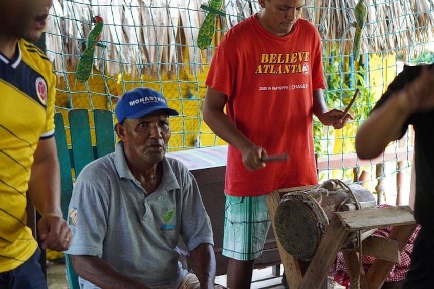 Un hombre con camisa roja y gorra azul trabajando con una herramienta, posiblemente tallando o cortando madera, con un fondo de malla y una estructura de madera cerca. 2