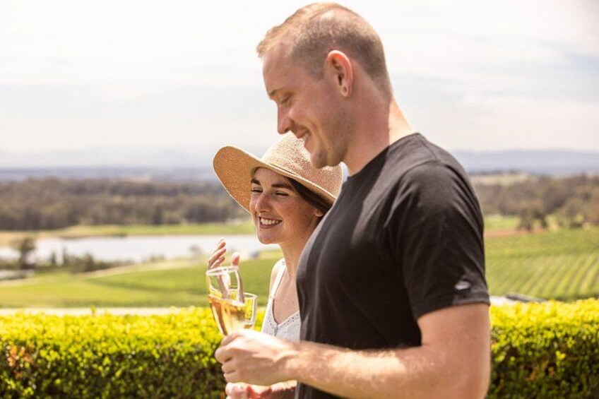 A person holding a glass of wine, another person wearing a hat, and a scenic view of a vineyard. 4