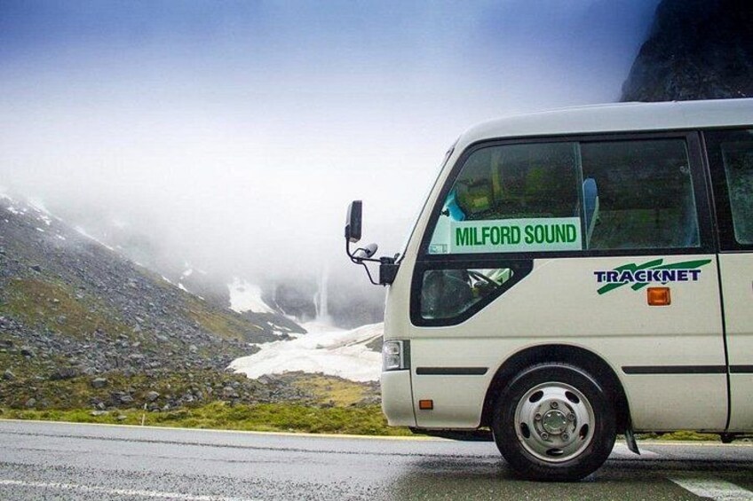 A white bus with 'MILFORD SOUND' and 'TRACKNET' on the side, parked on a road with a mountainous landscape and a waterfall in the background. 2