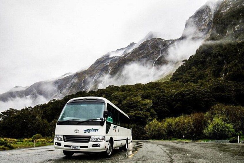 A white bus with 'TASMAN' written on the side, parked on a road with a mountainous landscape in the background. 3