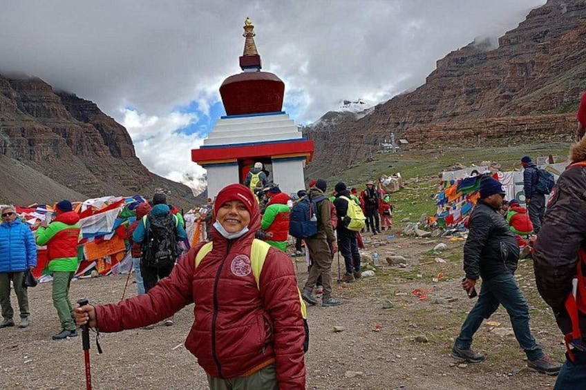A person in a red jacket holding a walking stick, a colorful prayer flag, and a group of people around a small shrine with a red and white structure. 4