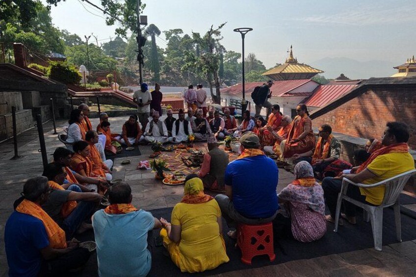 A group of people sitting in a circle outdoors, some in traditional attire, with a temple in the background. 5