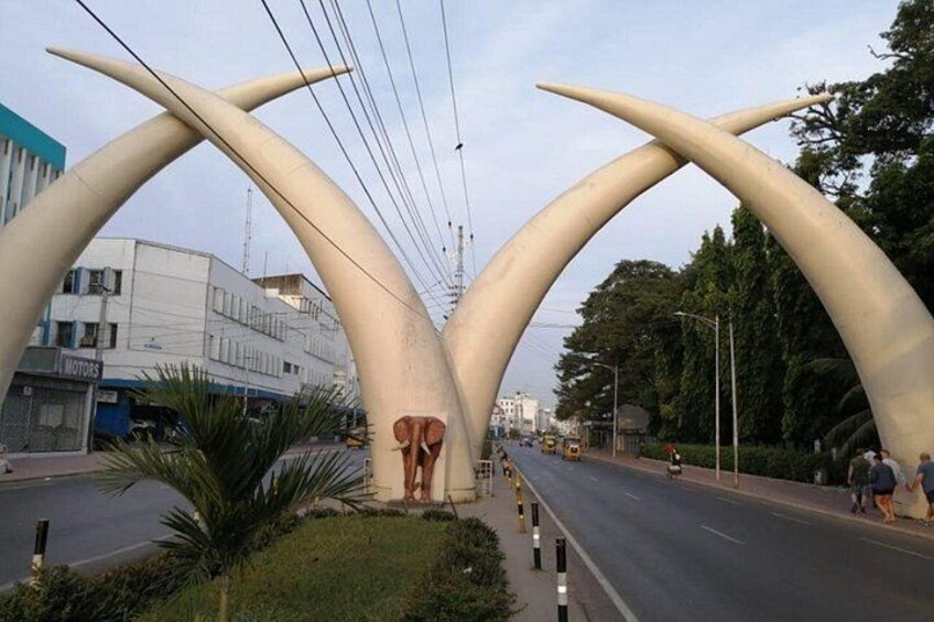 A road with a unique sculpture of a large, abstract figure in the center, surrounded by modern buildings and greenery. 1