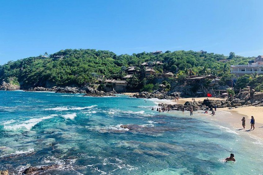 A beach with clear turquoise water, people swimming and walking on the shore, and a hill with buildings in the background. 1
