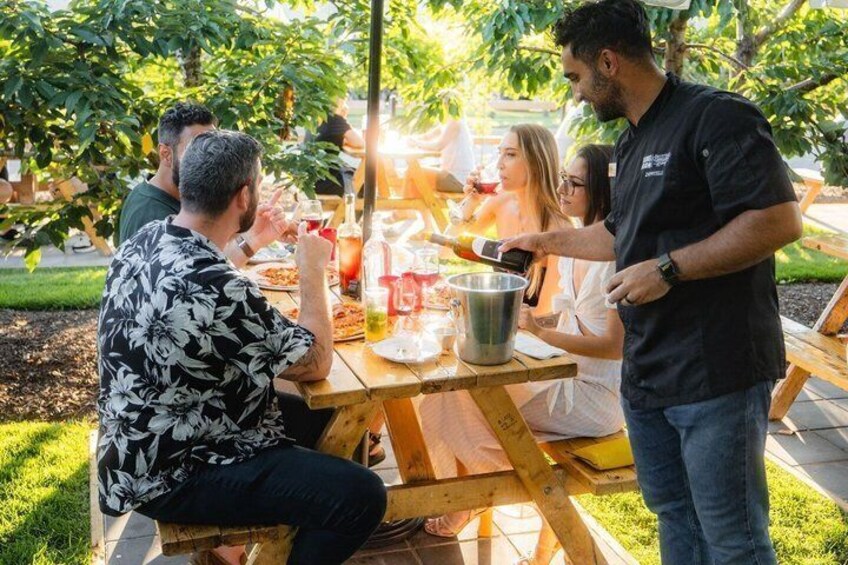 Personas sentadas en una mesa de madera al aire libre, con bebidas y comida, rodeadas de árboles. 1