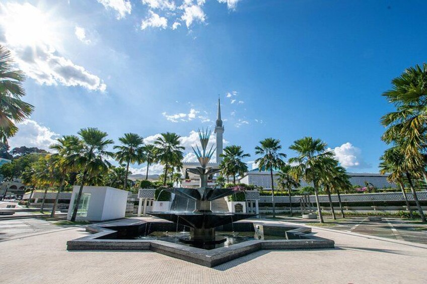 Una fuente moderna con un campanario, rodeada de palmeras y un cielo azul despejado. 3
