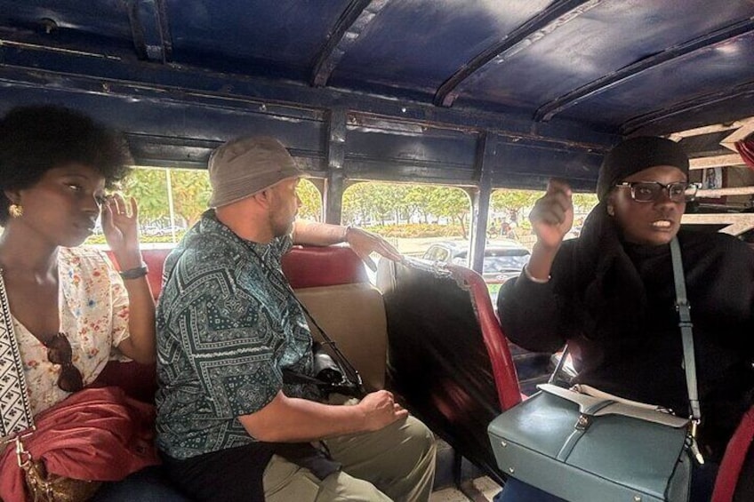Passagiere in einem Bus, ein Mann trägt ein gemustertes Hemd und eine Frau ein geblümtes Kleid. 4