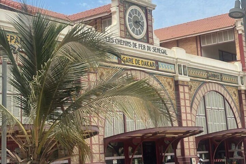 Ein Bahnhof mit einem Uhrturm, Palmen und Schildern mit der Aufschrift 'CHEMIN DE FER DU SENEGAL' und 'GAARE DE DAKAR'. 5