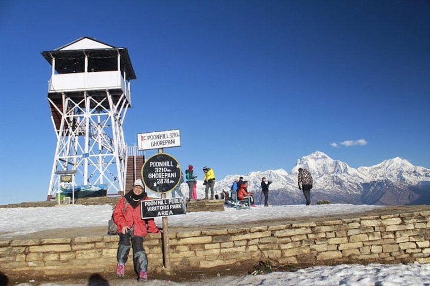 Montaña cubierta de nieve con una torre de madera para miradores, señales que indican Poon Hill y Ghorepanj, y personas haciendo senderismo. 1