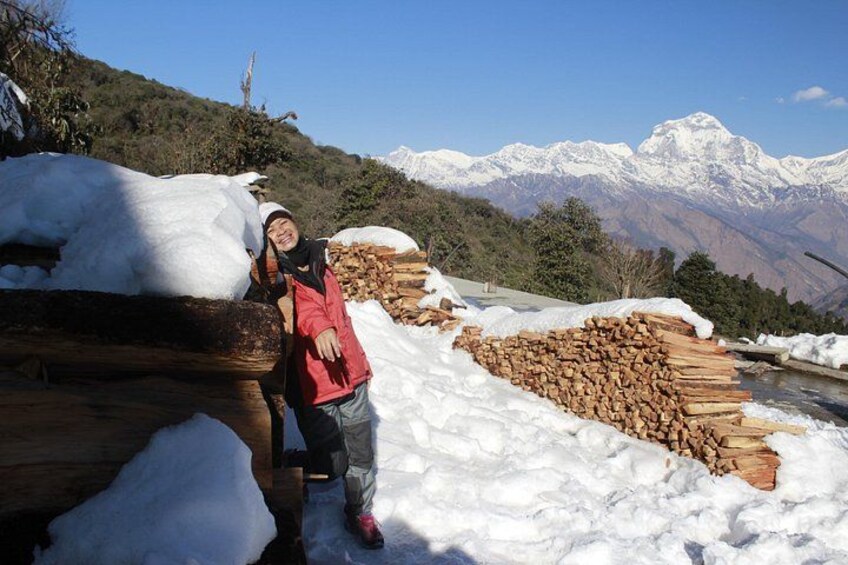 Una persona de pie junto a una pila de troncos y nieve en una zona montañosa con picos nevados al fondo. 3