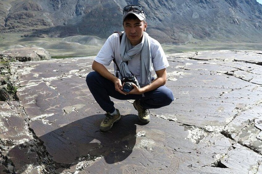 A person crouching on a rocky surface with a camera, wearing a cap, sunglasses, and a scarf. 4