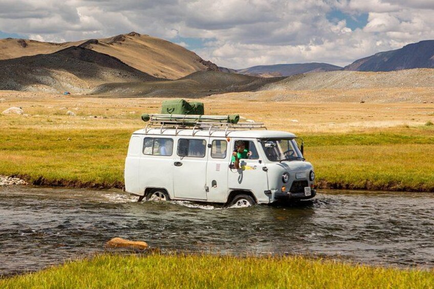 A white van driving through a shallow river in a grassy landscape with mountains in the background. 3