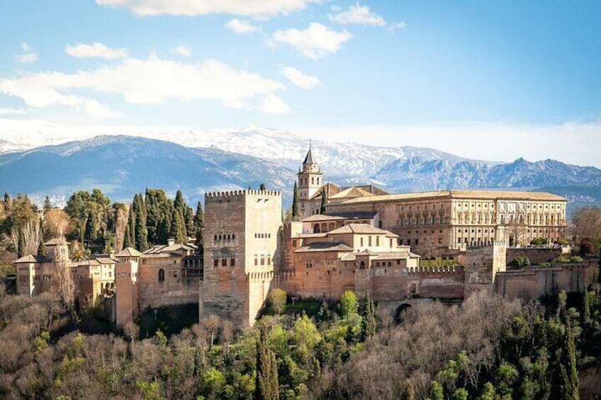 A historic castle complex with a prominent tower and a large building with a tiled roof, surrounded by trees and mountains in the background. 3
