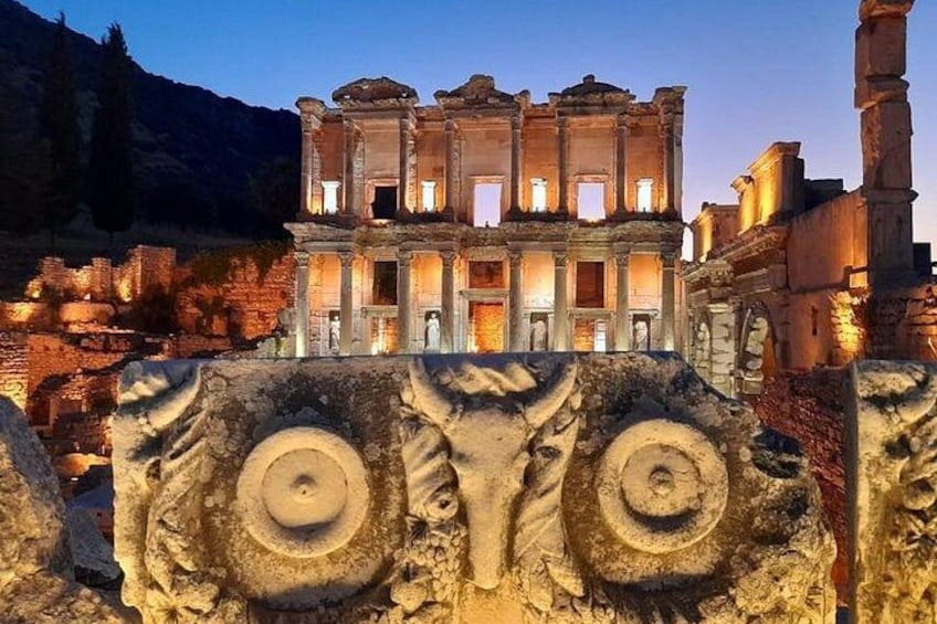 An ancient ruin with a large, illuminated structure, possibly a temple, surrounded by stone carvings and a mountainous backdrop. 1