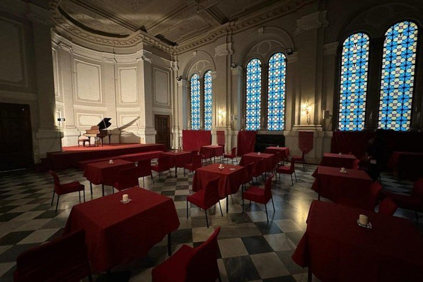 Un salón amplio con mesas y sillas rojas, un piano y grandes ventanales de vitral. 2