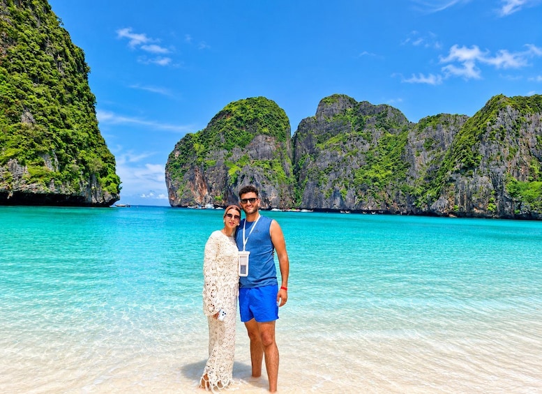 Un couple se tient sur une plage, avec une eau turquoise et limpide et de hautes falaises verdoyantes en arrière-plan. 4