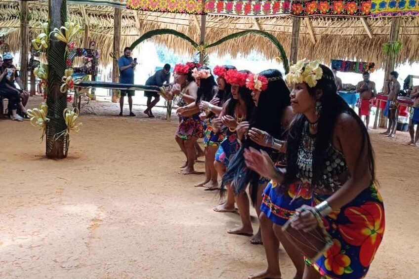 A group of people in colorful costumes, standing in a line under a thatched roof structure. 2